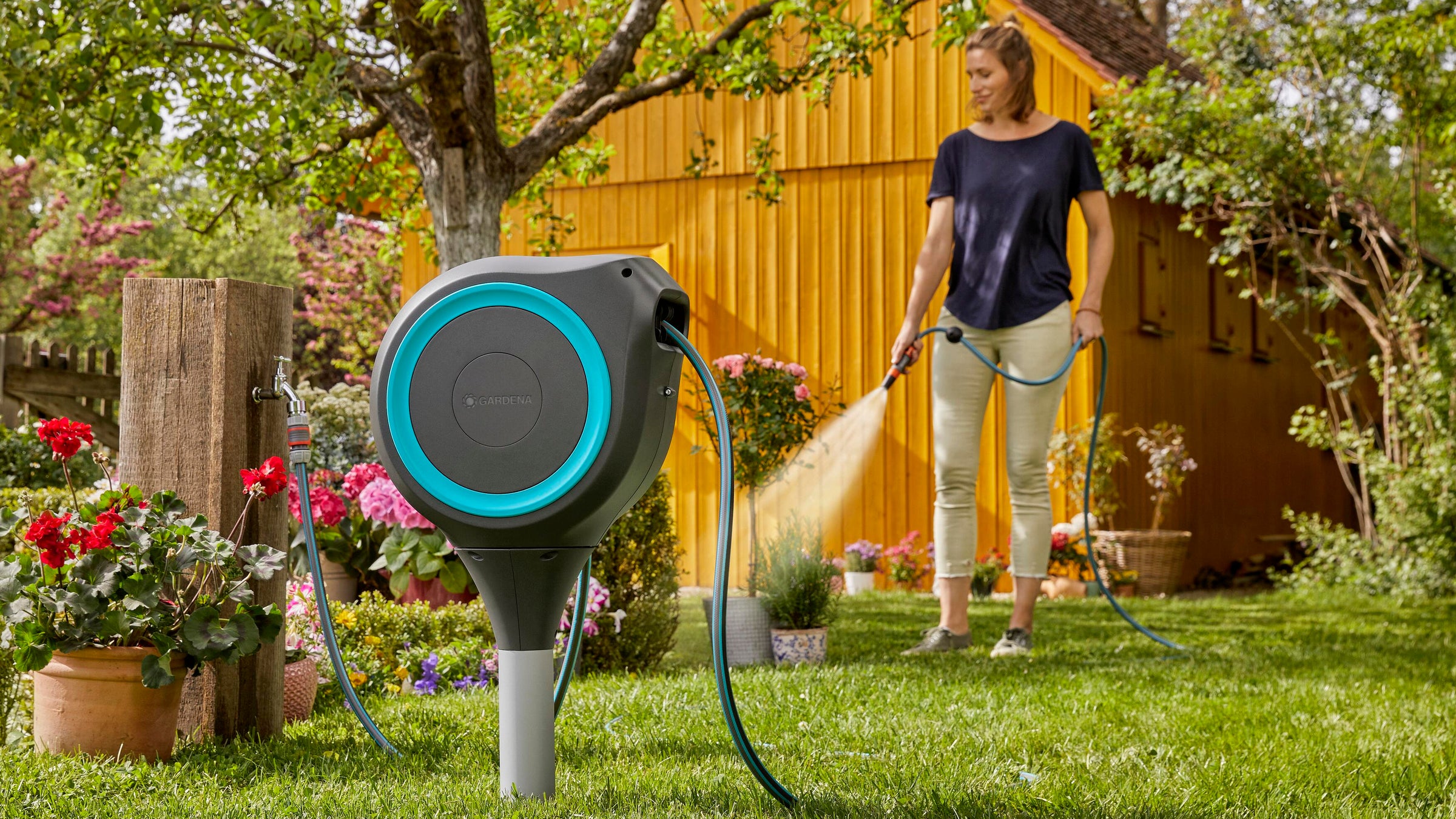 A woman watering a garden with a hose in front of a yellow wooden shed. In the foreground, a retractable garden hose reel with a black body and bright blue circular accent is mounted on a green lawn.